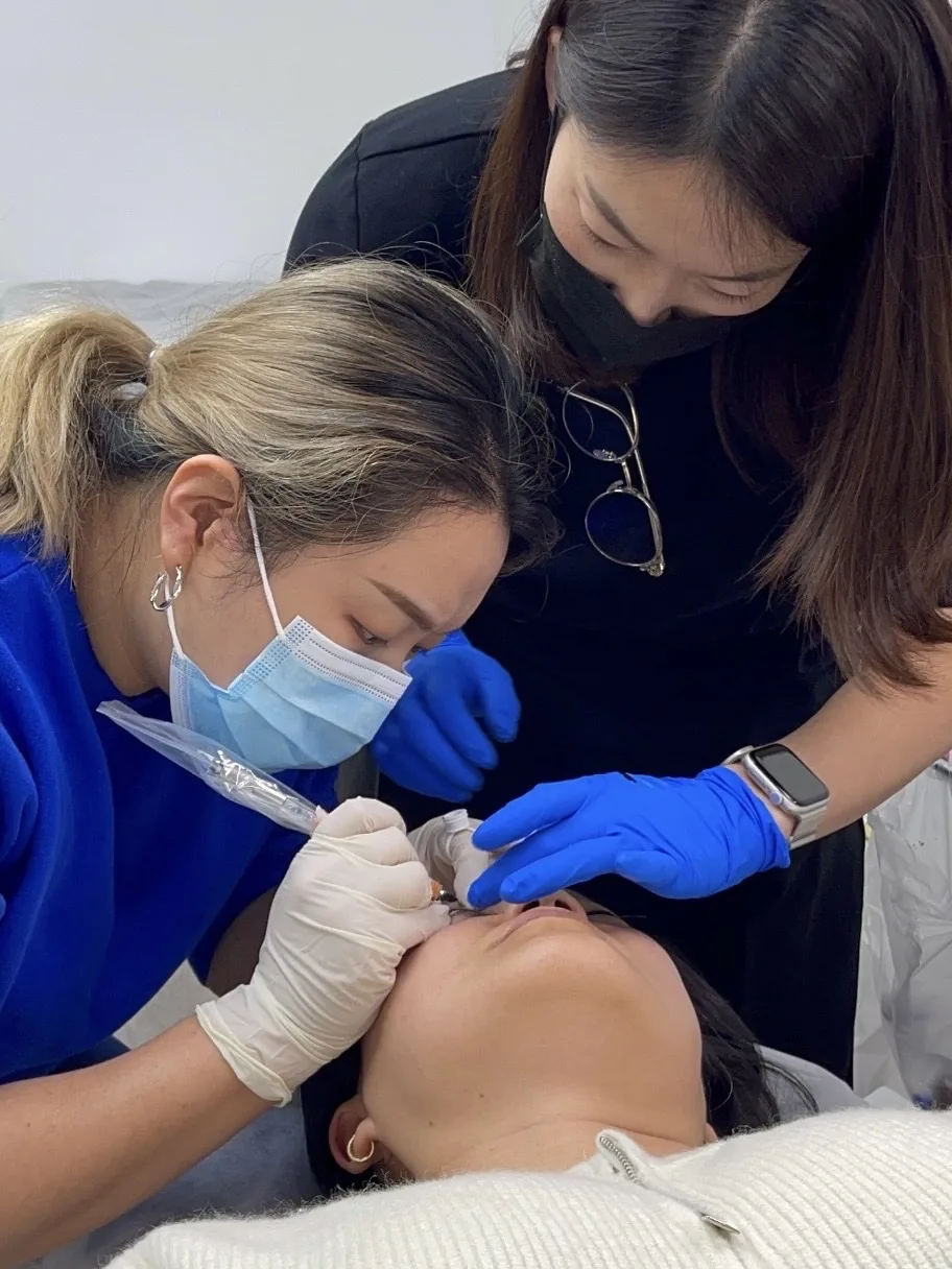 Student performing a permanent eyeliner procedure on a live model with close guidance from lead educator Sunny at SUNNYBROW Academy, Los Angeles