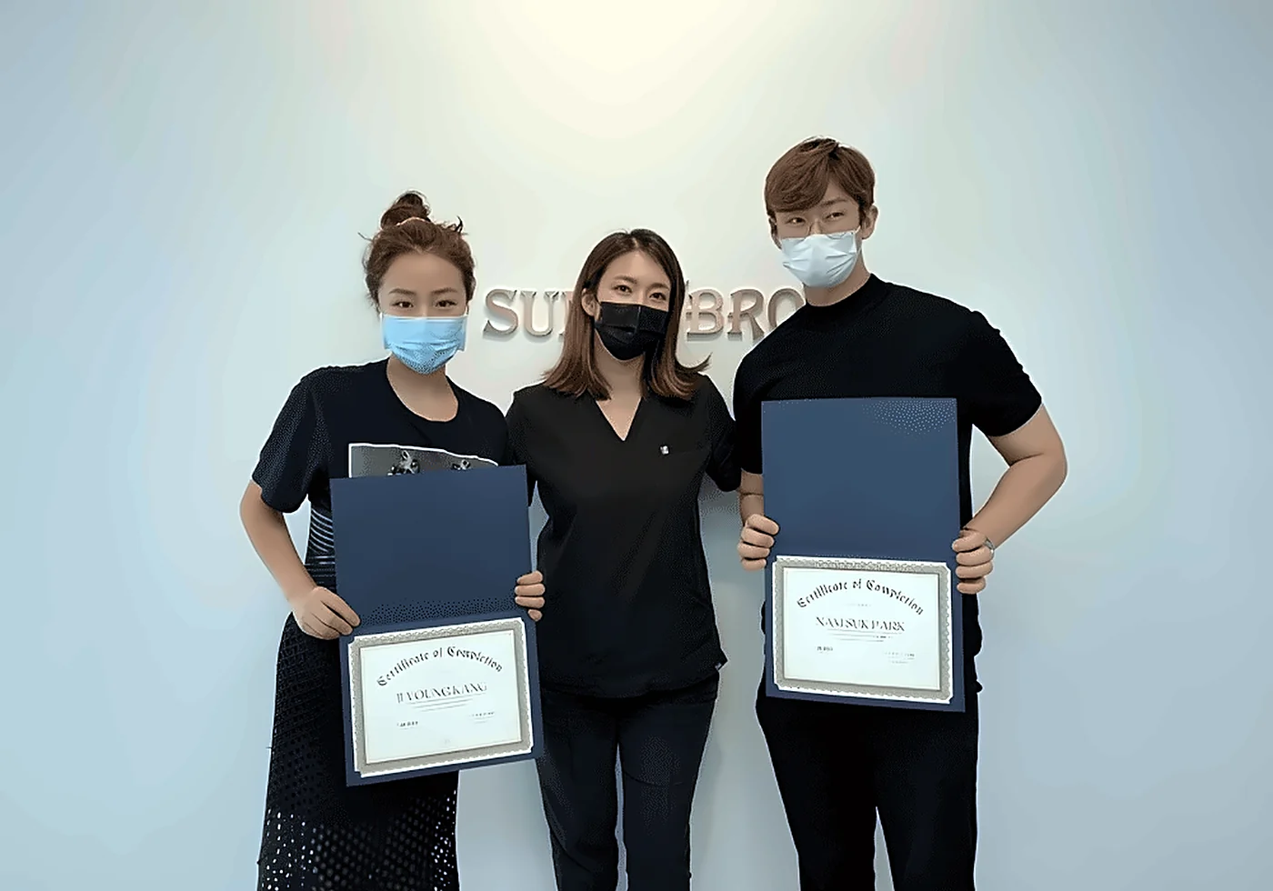 Two graduates holding their certificates of completion with lead educator Sunny at SUNNYBROW Academy, a permanent makeup school in Los Angeles